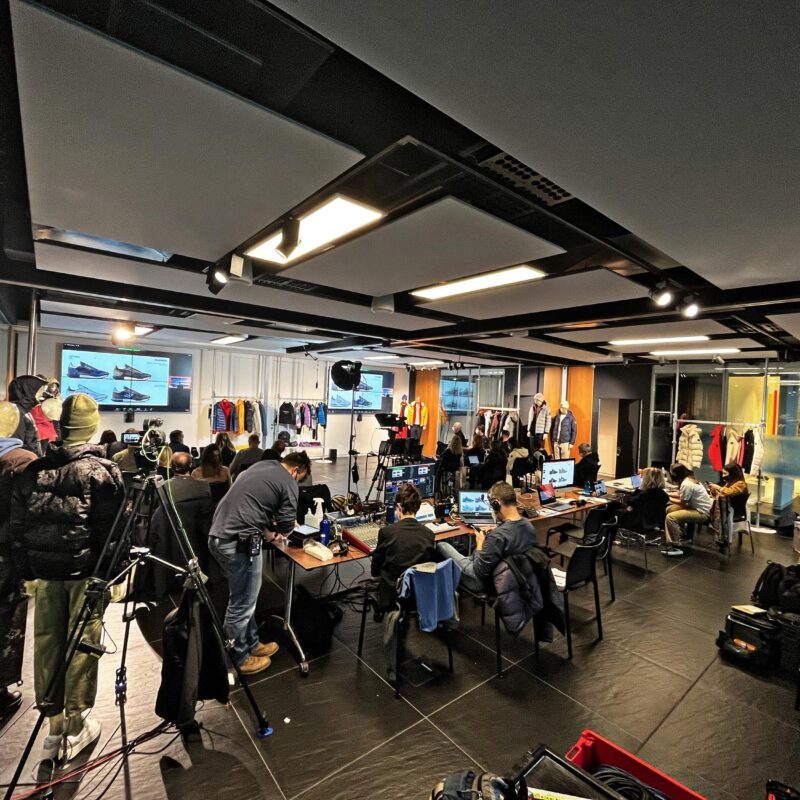 Uptelling live streaming production team at Napapijri Go To Market event — video and audio control desk with MacBooks, StreamDeck, audio mixer and video monitors, Napapijri showroom with large screen presentation, broadcast via Zoom